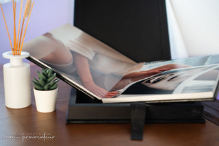 An open luxury boudoir photo album from Icon Provocateur displayed on a wooden table, featuring a soft-focus image of a woman in a white sweater and lace lingerie. The album rests in a black display box, styled with a small succulent and a white diffuser for a warm, intimate setting at Icon Provocateur