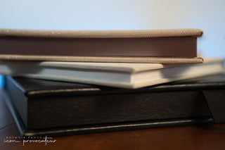 A close-up of a stack of luxury boudoir photo albums from Icon Provocateur, showcasing textured linen, shimmer fabric, and matte black wood covers. The image highlights the premium materials and craftsmanship available for clients’ printed artwork.