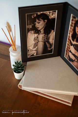 Close-up of a boudoir folio displaying a black and white portrait of a woman holding a coffee cup, alongside stacked linen-covered photo albums. The setup includes a small succulent and diffuser, emphasizing a luxurious and intimate presentation from Icon Provocateur.