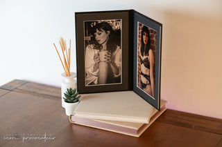 A matted folio display featuring two boudoir portraits—one of a woman in lingerie and one in a white shirt holding a cup, shown standing on two closed luxury albums atop a wooden table with soft decor. A product offering from Icon Provocateur.