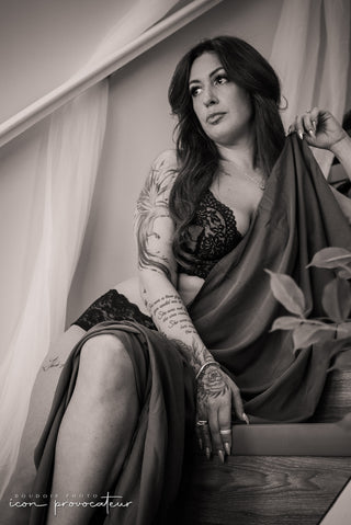 Boudoir portrait of a tattooed woman in black lace lingerie sitting gracefully on a staircase, draped in fabric, with soft natural light highlighting her serene expression.