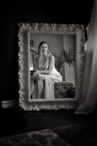 Bridal boudoir portrait of a woman in a satin robe, seated and gazing softly at her reflection in an ornate mirror, a timeless and elegant moment captured by Icon Provocateur, Montreal’s inclusive luxury studio.