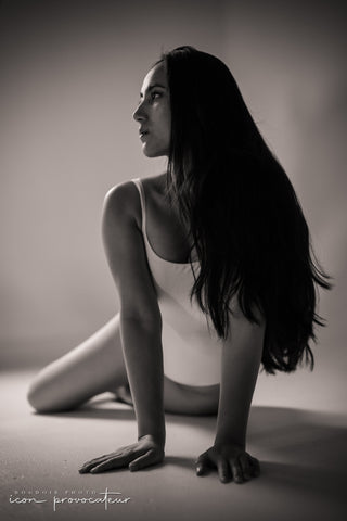 A woman in a white bodysuit sits on the floor with her arms extended, torso upright, and gaze directed to the side. Her long dark hair flows over one shoulder, and the soft black-and-white lighting emphasizes her graceful posture and the clean studio setting.