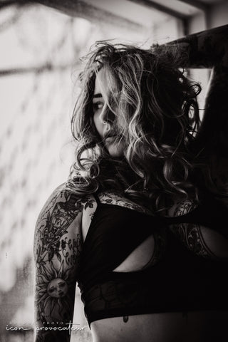 A tattooed woman stands in front of a window in a mesh cutout top, her tousled hair partially obscuring her face. Captured in dramatic black and white for the Icon experience at Icon Provocateur, embodying bold, rebellious femininity.