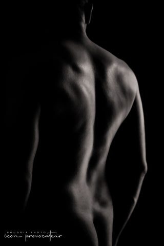 An artistic black and white photograph of a man's nude back, with dramatic side lighting revealing the muscular contours of his shoulders and spine. The image emphasizes strength and form through light and shadow.