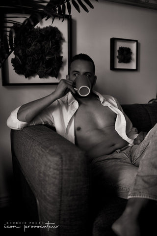 A man lounges shirtless on a sofa with an open white button-down and jeans, sipping from a mug. Soft shadows and a neutral palette create a relaxed, masculine atmosphere in this lifestyle-inspired boudoir image.