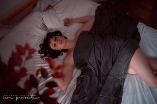 Boudoir portrait of a woman lying on a bed partially covered by dark sheets, photographed from above, cinematic and intimate image by Icon Provocateur, Montreal’s inclusive luxury studio.