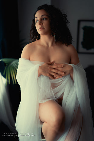 Boudoir portrait of a woman seated in soft white draping, hands crossed, gazing off-camera, poised and graceful imagery by Icon Provocateur, Montreal’s inclusive luxury studio.