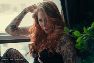 Close-up boudoir portrait of a tattooed woman with long red hair, seated by a sunlit window — raw and confident imagery by Icon Provocateur, Montreal’s inclusive luxury studio.