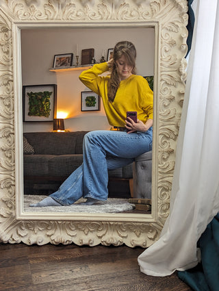 A behind-the-scenes mirror selfie of Gal at the Icon Provocateur studio, wearing a yellow sweater and jeans, with cozy decor and framed greenery in the background.