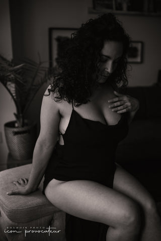 Black and white boudoir portrait of a woman seated in a black dress, gazing downward in soft natural light, intimate and thoughtful image from the Flirt experience at Icon Provocateur, Montreal’s inclusive luxury studio.