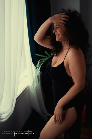 Boudoir portrait of a woman laughing by the window in soft natural light, wearing a black bodysuit, joyful and relaxed image from the Flirt experience at Icon Provocateur, Montreal’s inclusive luxury studio.