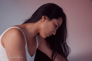 A woman in a white bodysuit sits in profile with her head bowed, her long black hair cascading forward. The soft lighting and minimal pink-to-purple gradient backdrop create a serene, introspective mood in this Icon Provocateur boudoir portrait.