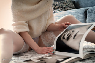 Close-up of a woman in lace lingerie and a knit sweater browsing an art book while seated on the floor in soft natural light.