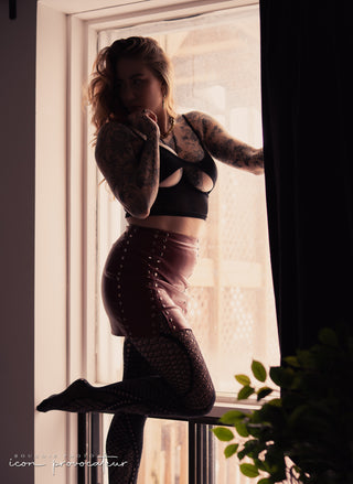 Tattooed woman in a leather skirt and lingerie poised sensually by a backlit window, an edgy, artful boudoir portrait by Icon Provocateur, Montreal’s inclusive luxury studio.