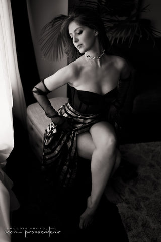 Elegant woman in corset and pearl choker seated near a window, one leg raised, her posture poised and statuesque, a sensual and sculptural boudoir portrait by Icon Provocateur, Montreal’s inclusive luxury studio