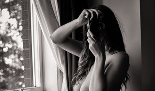 Black and white boudoir portrait of a topless woman holding a camera in front of her face, standing by a window with soft natural backlight, an intimate and artistic moment captured at Icon Provocateur, Montreal.