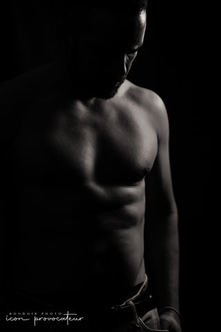 A shirtless man stands in dramatic shadow, his face partially obscured and his torso subtly lit to highlight muscle definition and texture. Wearing low-slung jeans, the image captures quiet strength and raw intimacy in a fine art boudoir style.
